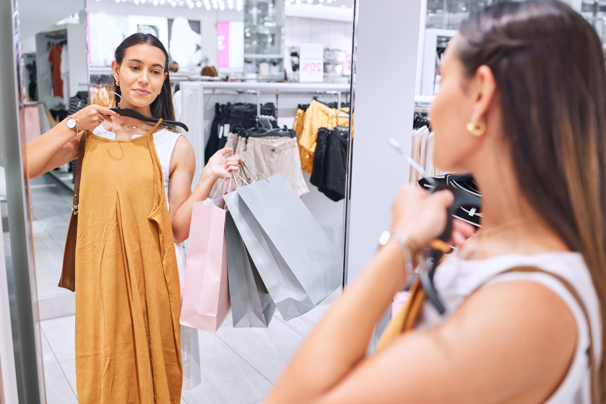 A woman holding shopping bags is trying on a mustard dress in front of a mirror inside a clothing store