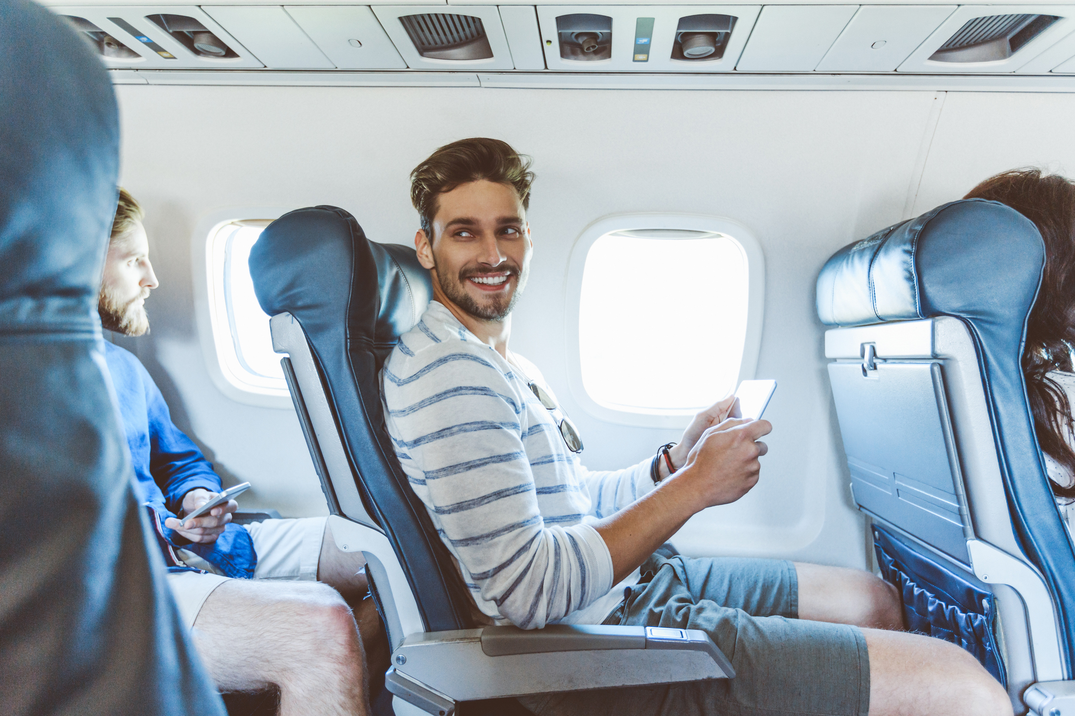 A smiling man in casual attire with a striped shirt and shorts holds a smartphone while sitting in an airplane seat. Another person is seen in the background