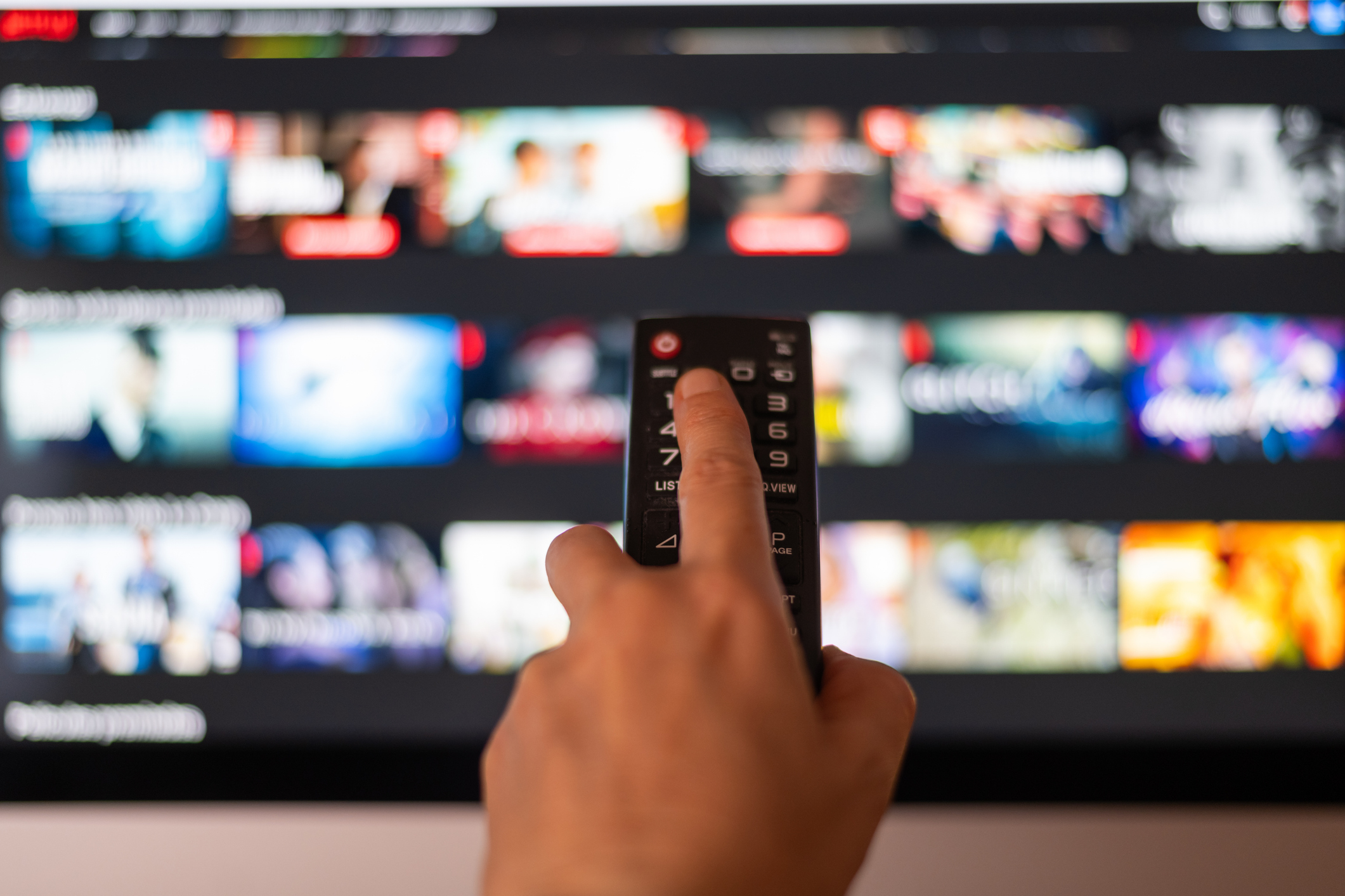 A hand holding a remote control points at a TV screen displaying various media thumbnails on a content streaming service