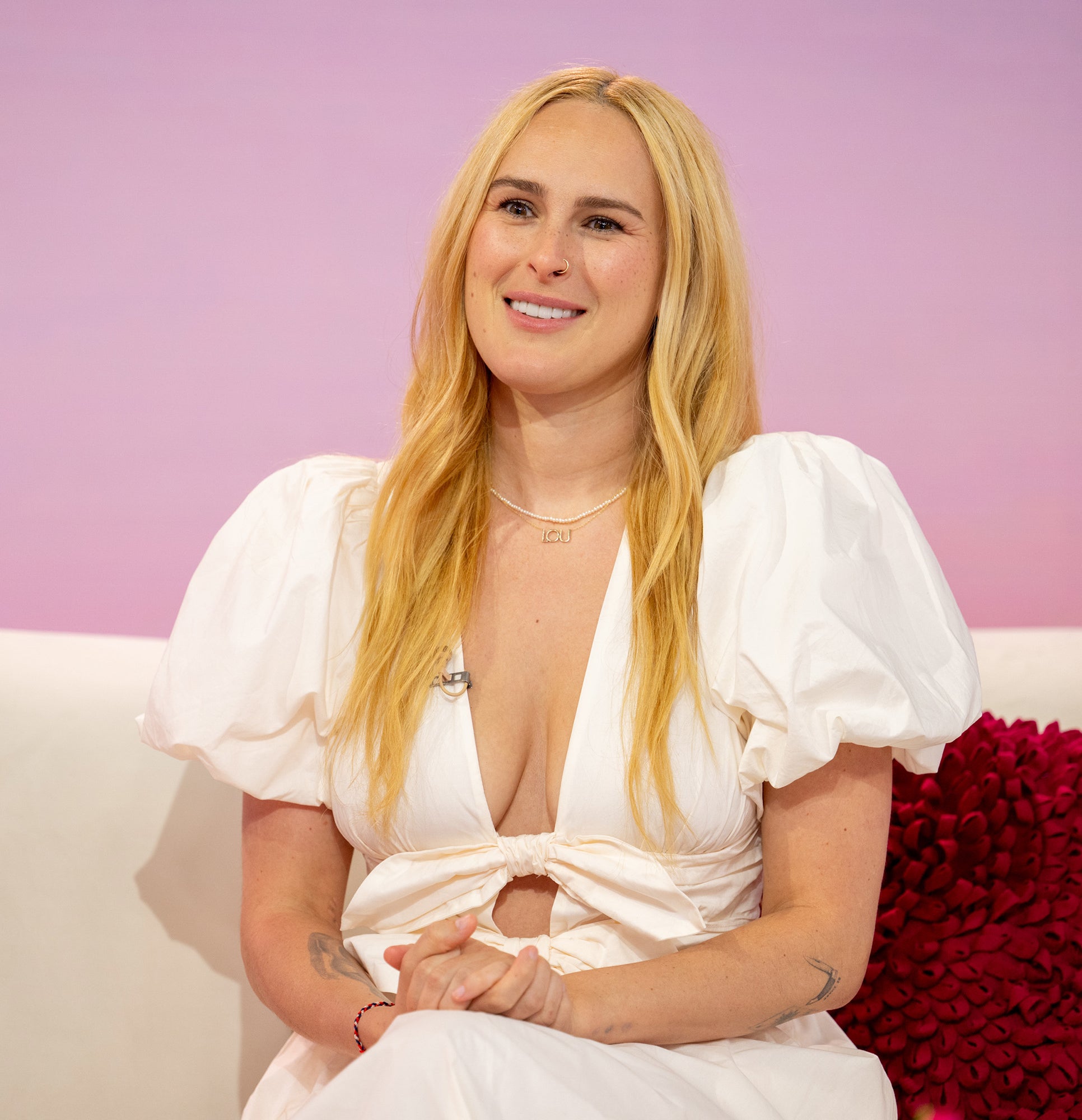 Rumer Willis sits on a couch wearing a white dress with puffed sleeves. She smiles with her hands clasped in her lap