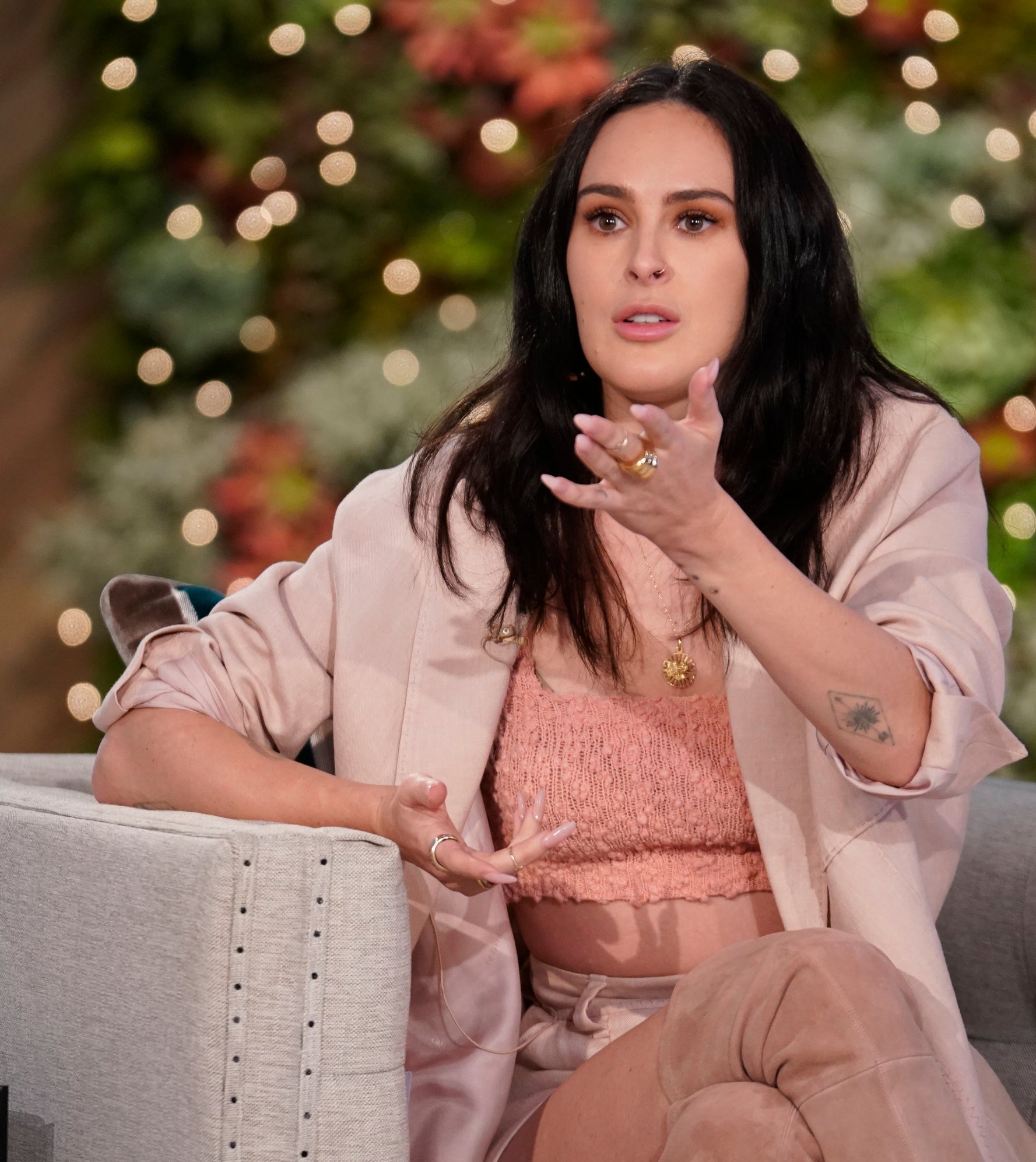 Rumer Willis is seated on a talk show set, gesturing while speaking. She wears a light-colored blazer and pants with a top underneath. A mug with &quot;The Talk&quot; logo is on the table