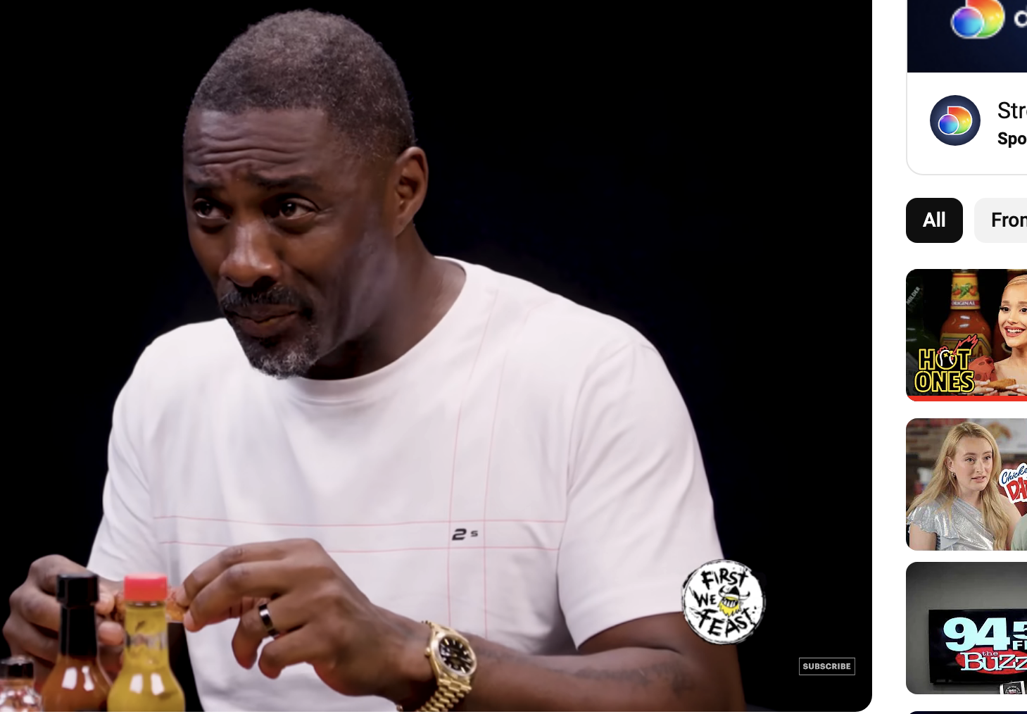 Idris Elba is seated at a table holding a hot sauce bottle on the YouTube show &quot;Hot Ones&quot; with bottles of hot sauce in front of him