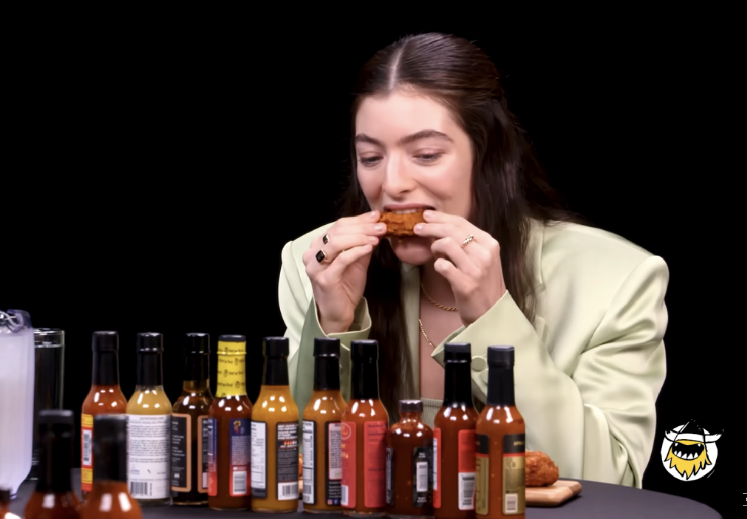 Lorde eating spicy wings on "Hot Ones" show, with various hot sauces on the table. Screenshot from YouTube titled "Lorde Drops the Mic While Eating Spicy Wings | Hot Ones"