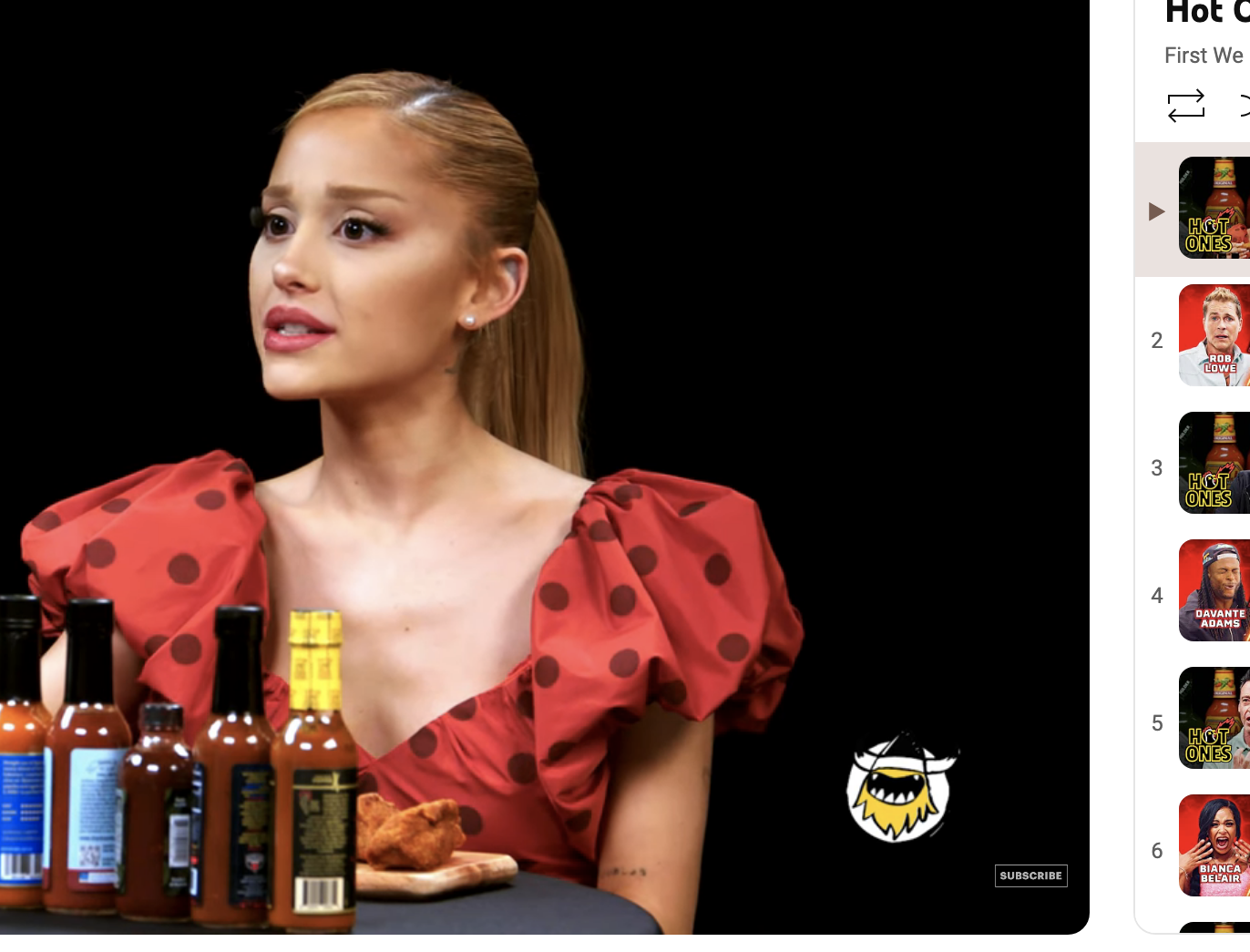 Ariana Grande on &quot;Hot Ones,&quot; a web series, in the interview setting with bottles of hot sauces on the table. She wears a polka-dot dress with puffed sleeves