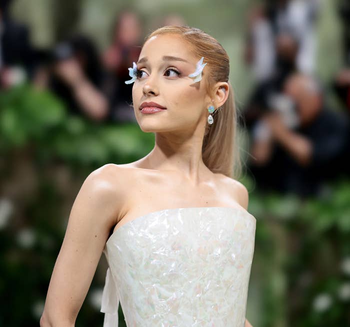 Ariana Grande is pictured on a red carpet in an elegant strapless dress with unique eye makeup featuring white designs extending from her eyes