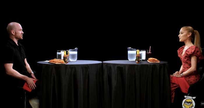 Ariana Grande and Sean Evans sit at a table with hot wings and drinks during a Hot Ones interview
