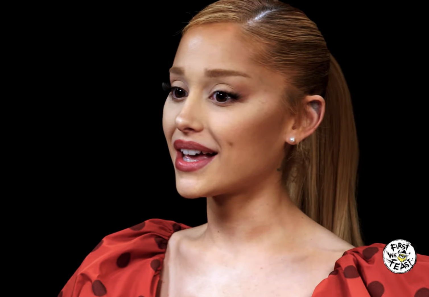 Ariana Grande on Hot Ones, talking while eating spicy wings. She is wearing a dress with puffed sleeves. The YouTube video page shows related videos on the right