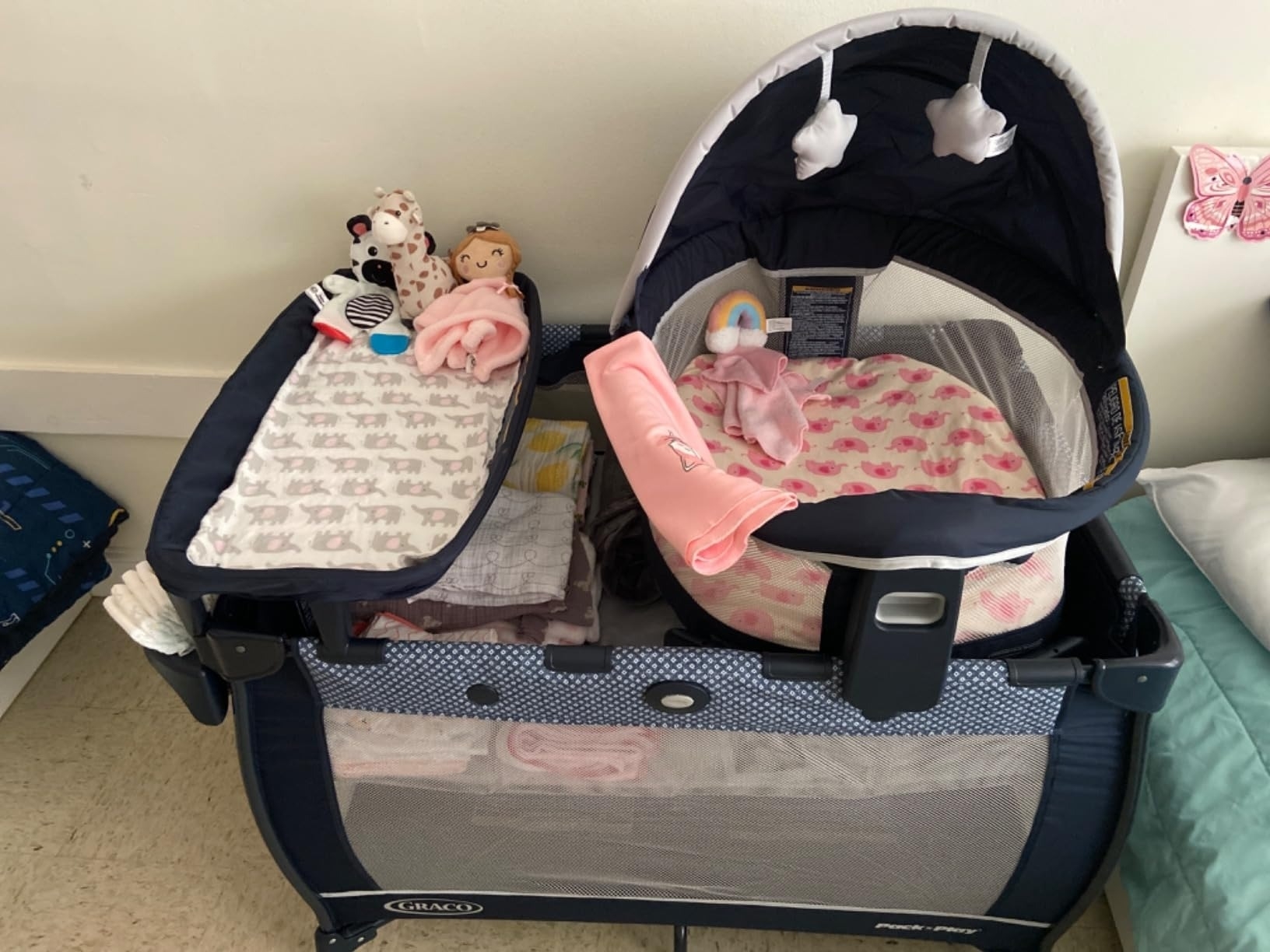 A Graco Pack 'n Play playard set up with a bassinet insert. It holds a plush blanket, a swaddle, a pacifier, and several stuffed toys
