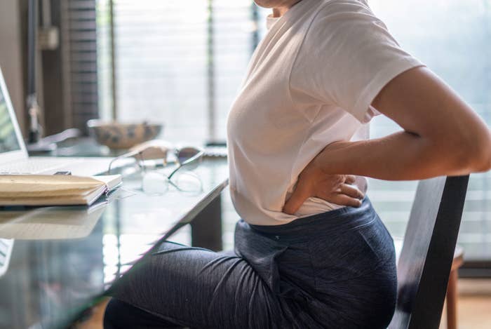 Person sitting at a desk, holding their lower back in pain, with a laptop, a pair of glasses, and an open notebook in the background