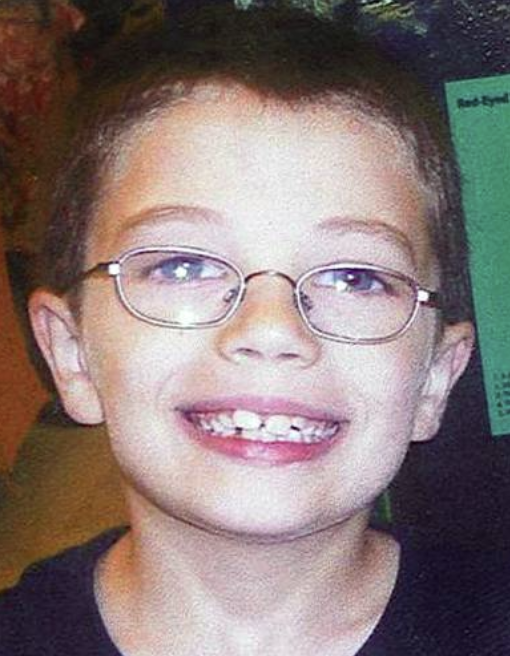 A young child with short hair and glasses, smiling with their teeth showing