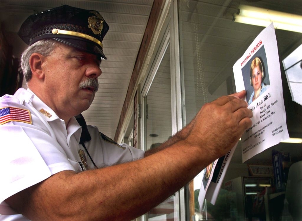 A police officer posts a missing person flyer of Molly Ann Bish on a shop window. The officer's uniform includes a cap and badge