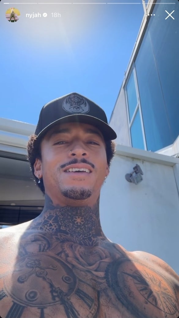 Nyjah Huston takes a selfie outside, shirtless, showing his tattooed chest and wearing a black cap. The sky is clear and a building is partially visible