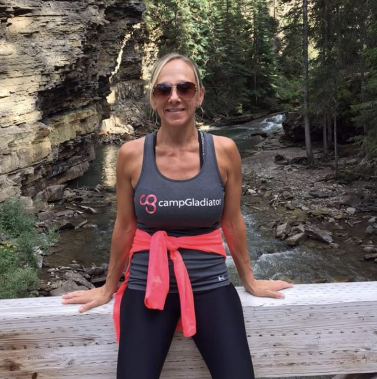 A woman wearing a "campGladiator" tank top and sunglasses is smiling and posing on a wooden railing in front of a rocky river landscape