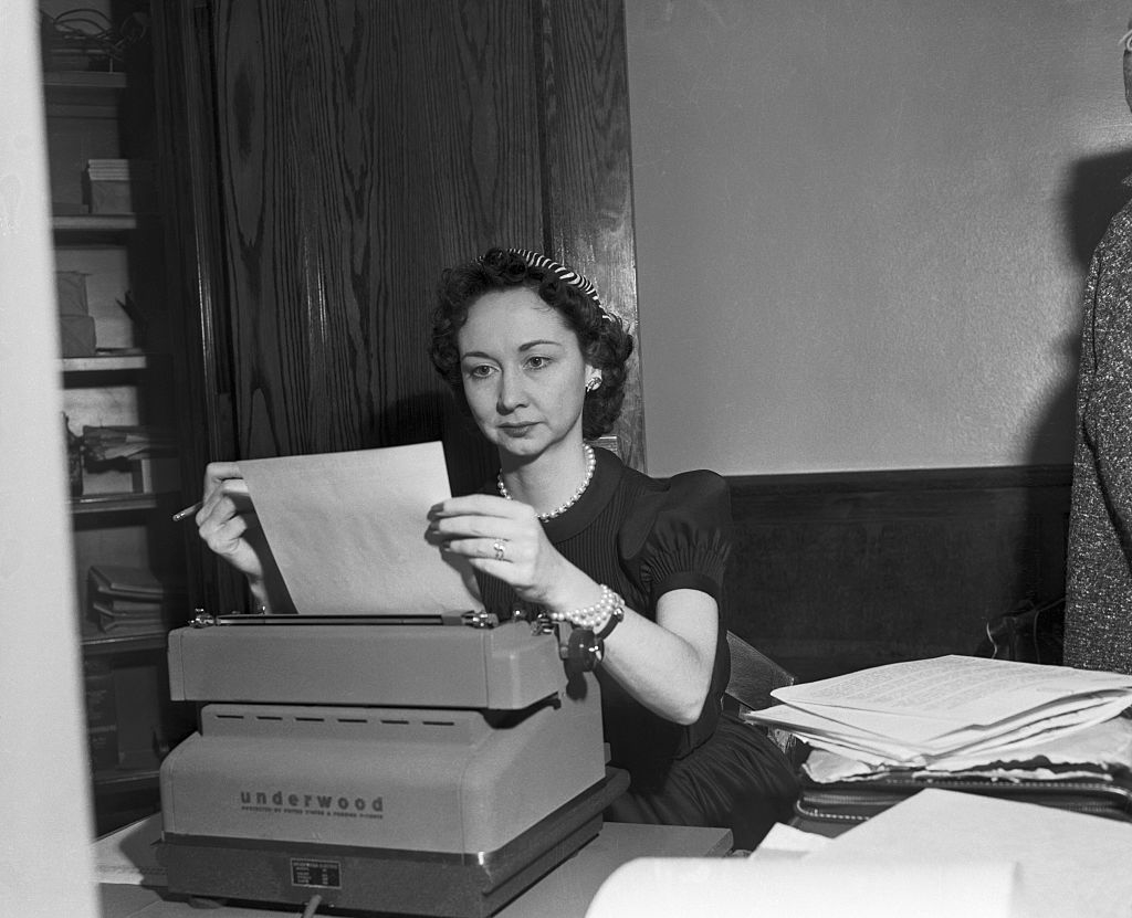A woman sits at a desk using an Underwood typewriter, focusing intently on her work