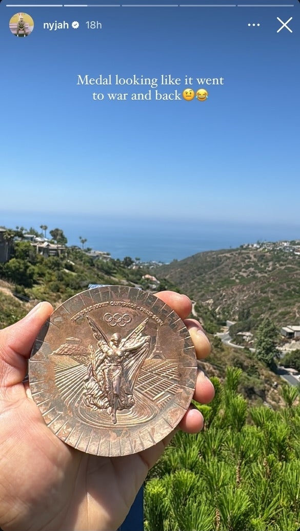 Close-up of a hand holding a worn Olympic medal with a scenic background of hills and the ocean. Text overlay reads, "Medal looking like it went to war and back" with emojis