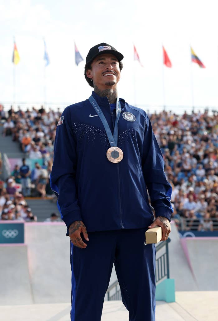 Nyjah Huston stands proudly with a bronze medal around his neck, dressed in a blue athletic tracksuit and cap at the Olympic Games with spectators in the background