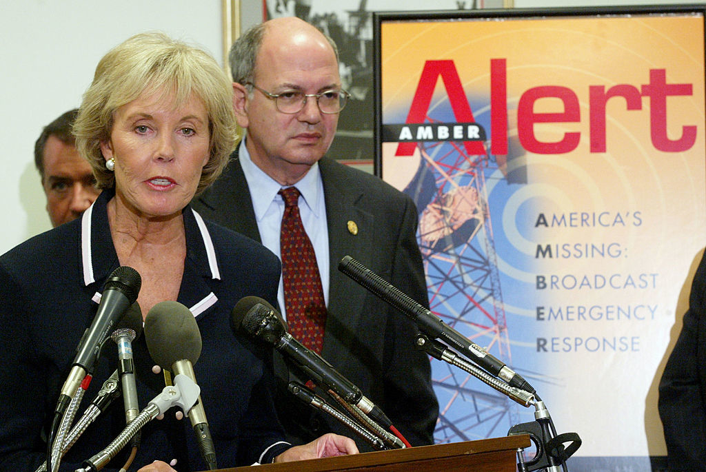 Kim Gandy speaks at a press conference in front of a sign that says "Amber Alert: America's Missing: Broadcast Emergency Response" while an unidentified man stands behind her