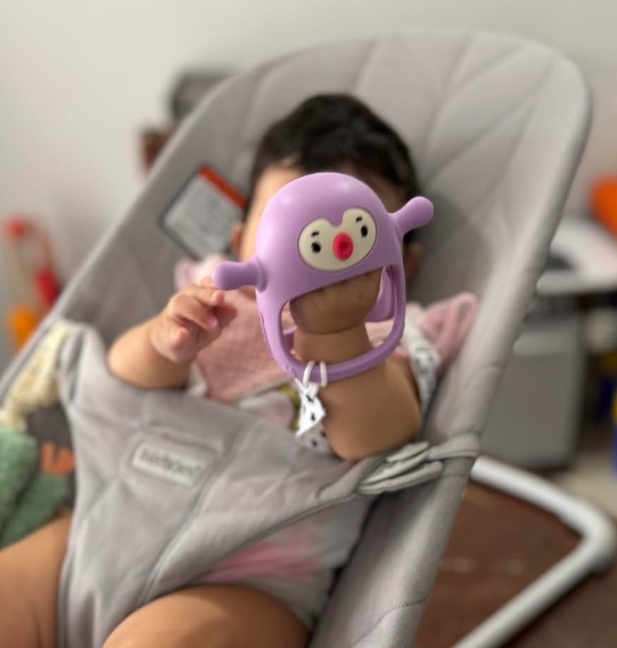 A baby sits in a bouncer holding a purple toy with a face. Various toys are visible in the background