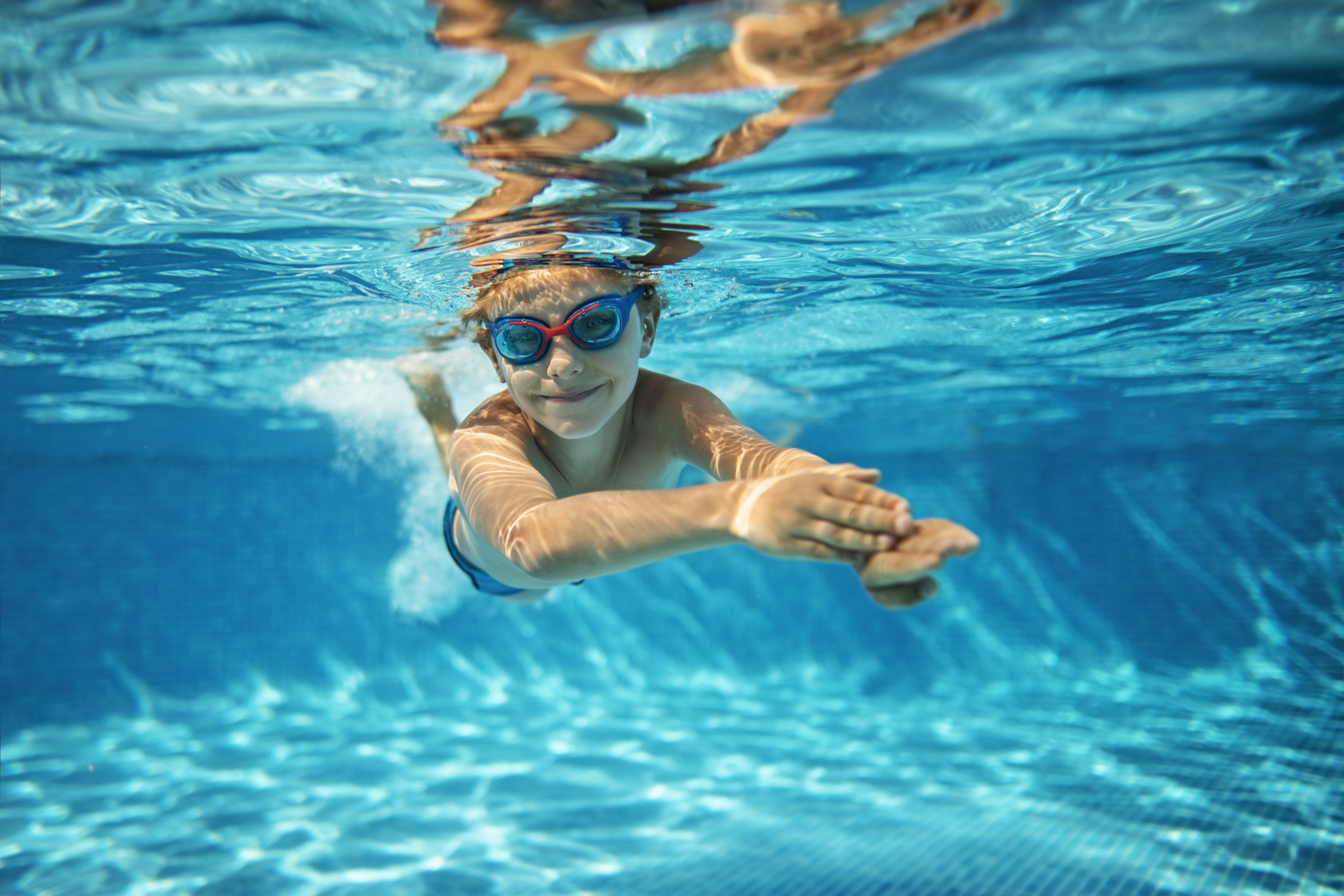 A young child with swim goggles swims underwater in a pool, smiling and reaching forward with their hands