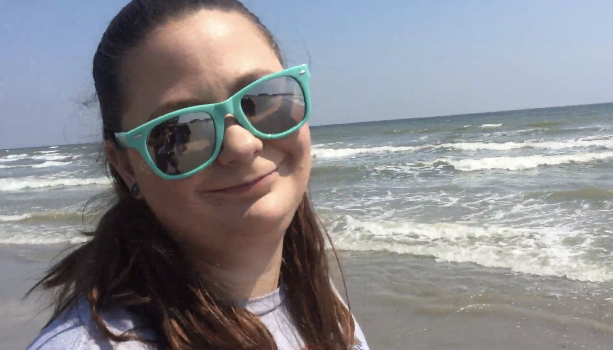 A woman with long hair, wearing sunglasses, smiles slightly while standing on the beach with waves in the background