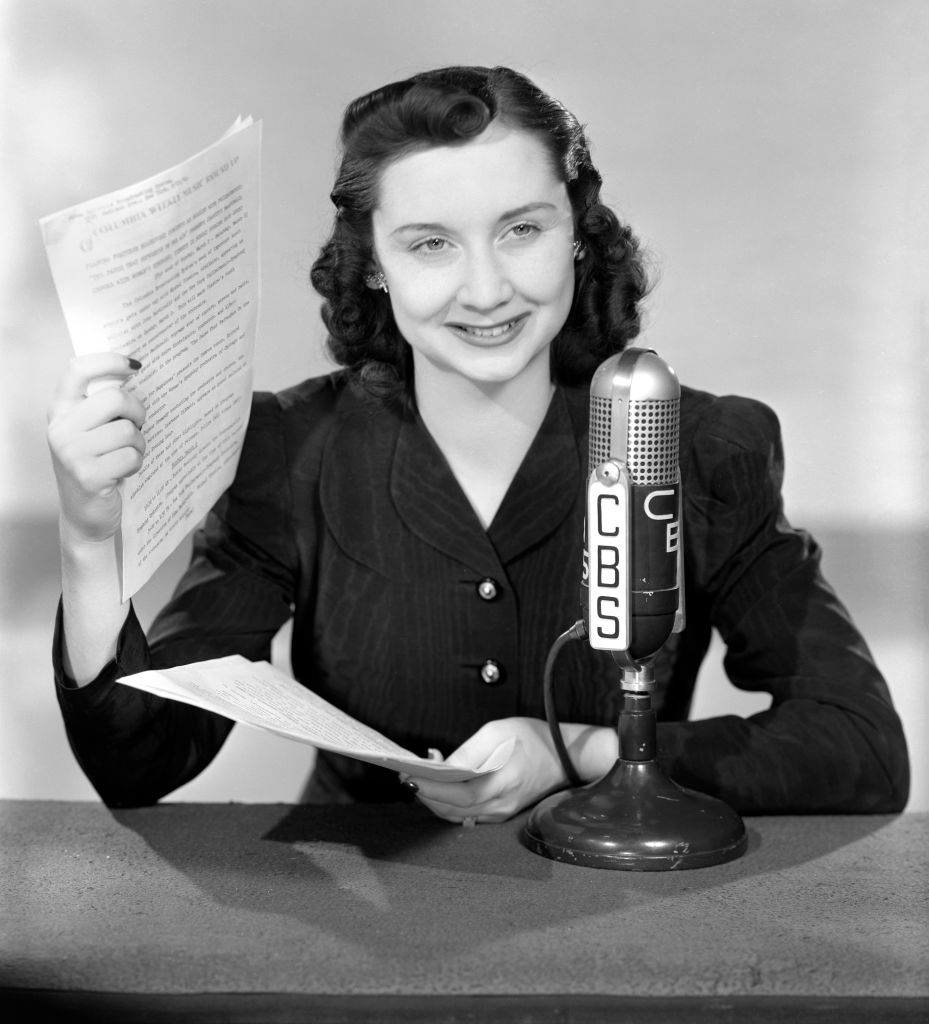 A woman in vintage attire, seated at a desk, holds papers while speaking into an old CBS microphone