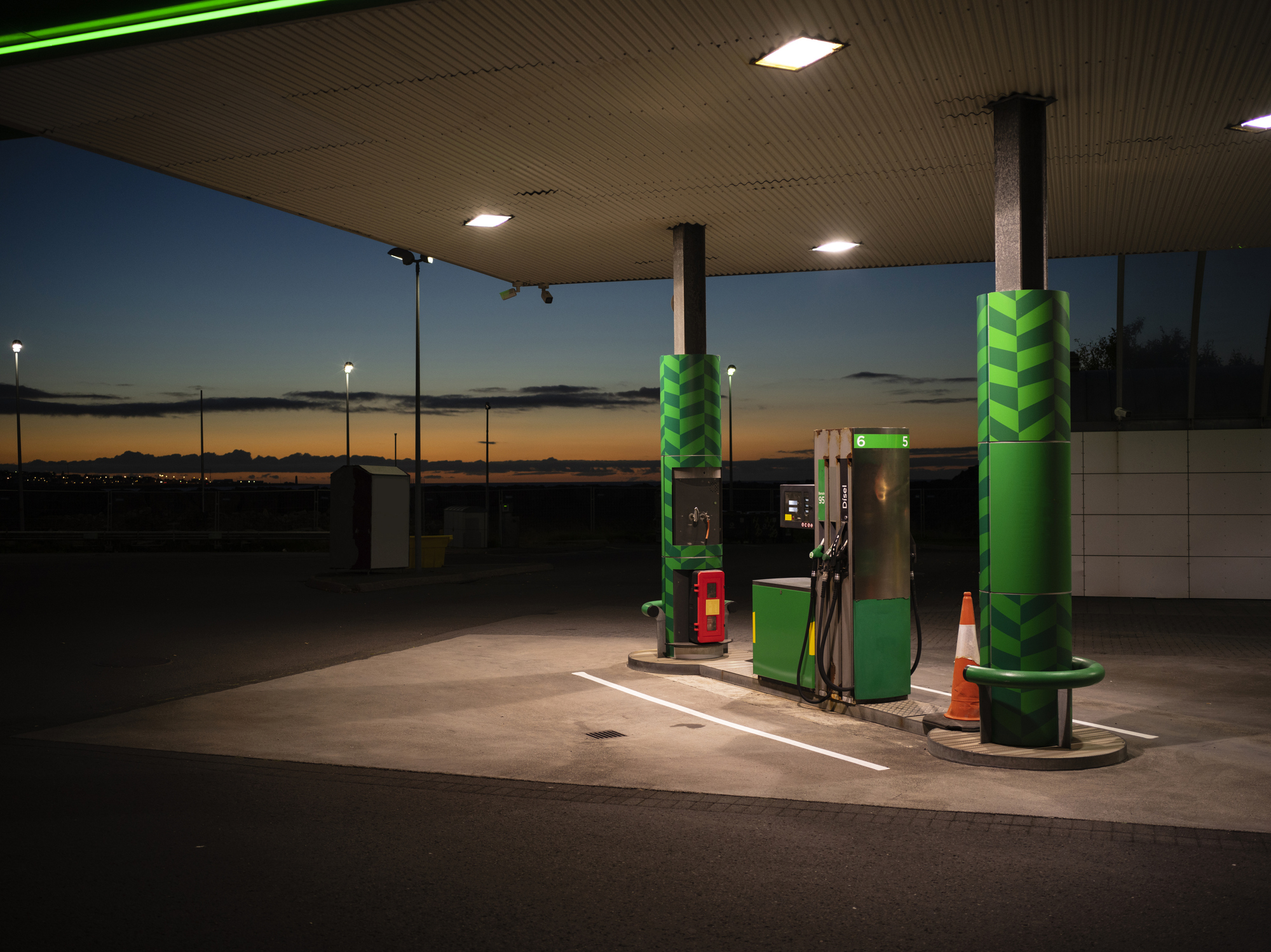 A gas station at twilight with lit overhead lights, green pillars, and two fuel pumps. The surrounding area appears empty and quiet