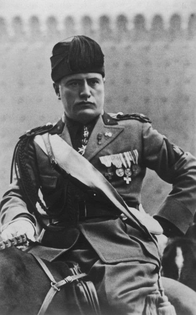 A historical black and white photograph of Benito Mussolini in a military uniform, seated on a horse. The background shows a blurred structure