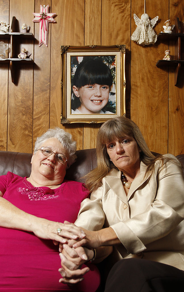 An older woman and a middle-aged woman hold hands on a couch, with a framed photo of a young girl on the wall behind them