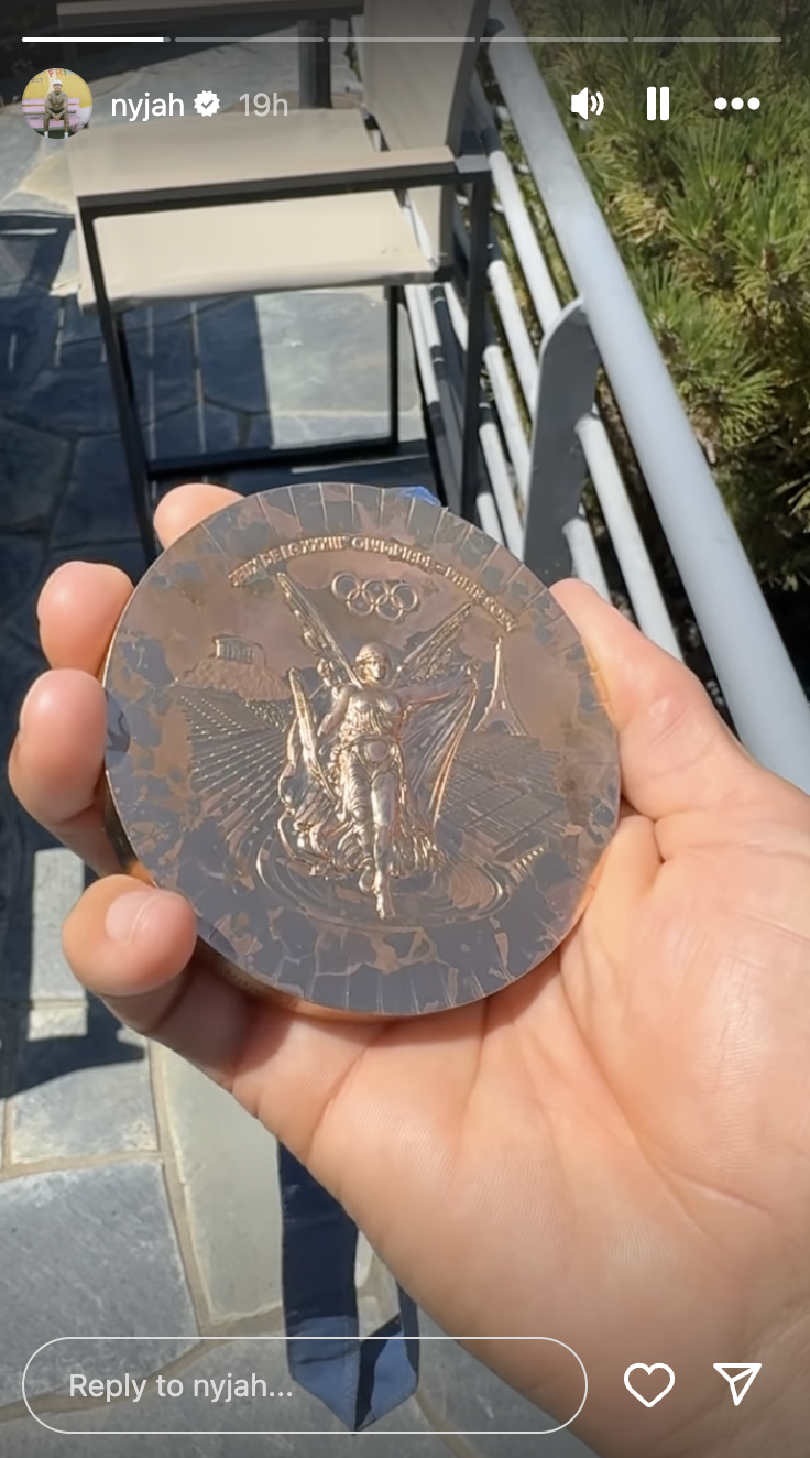 A close-up of a hand holding an Olympic medal. The medal features a raised design of an ancient Olympic scene