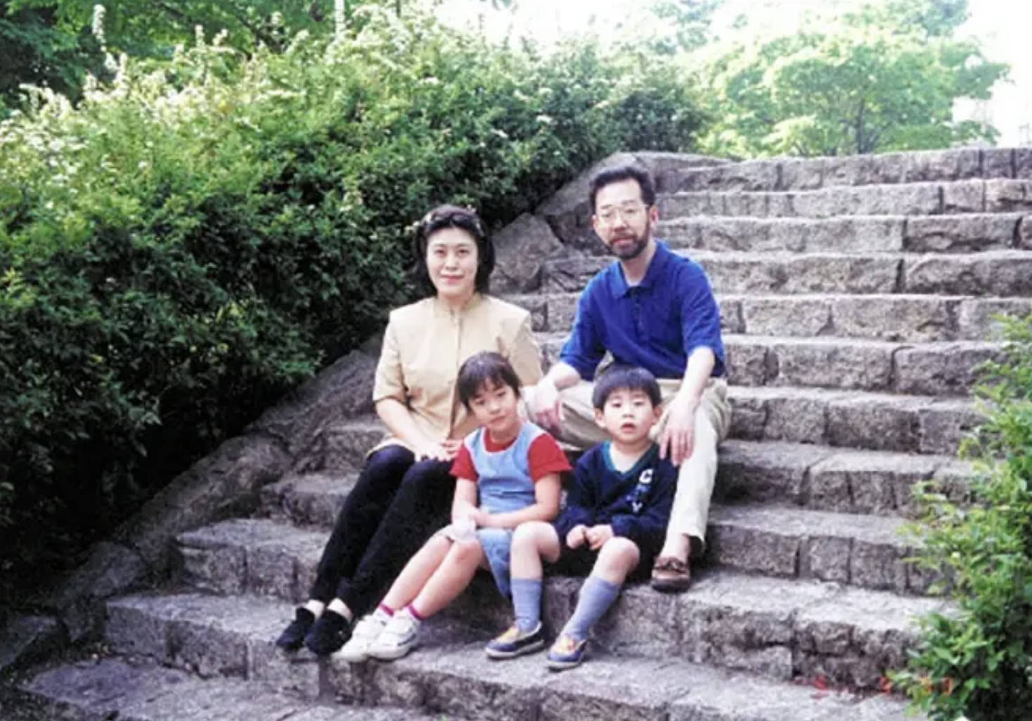 A family of four sits on stone steps outdoors. The parents, a man and a woman, sit with their two children, a boy and a girl. Green foliage surrounds the steps