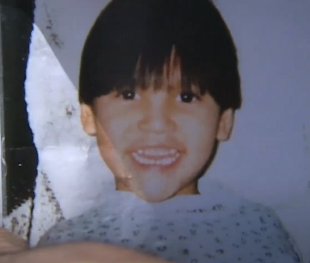 A young child smiles brightly at the camera, revealing their baby teeth. The child has dark hair with bangs covering their forehead and is wearing a light-colored top