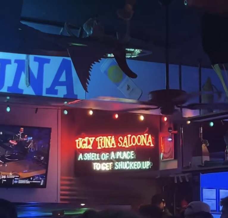 Crowded scene inside Ugly Tuna Saloona with a neon sign reading "A shell of a place, to get shucked up!" above the bar area