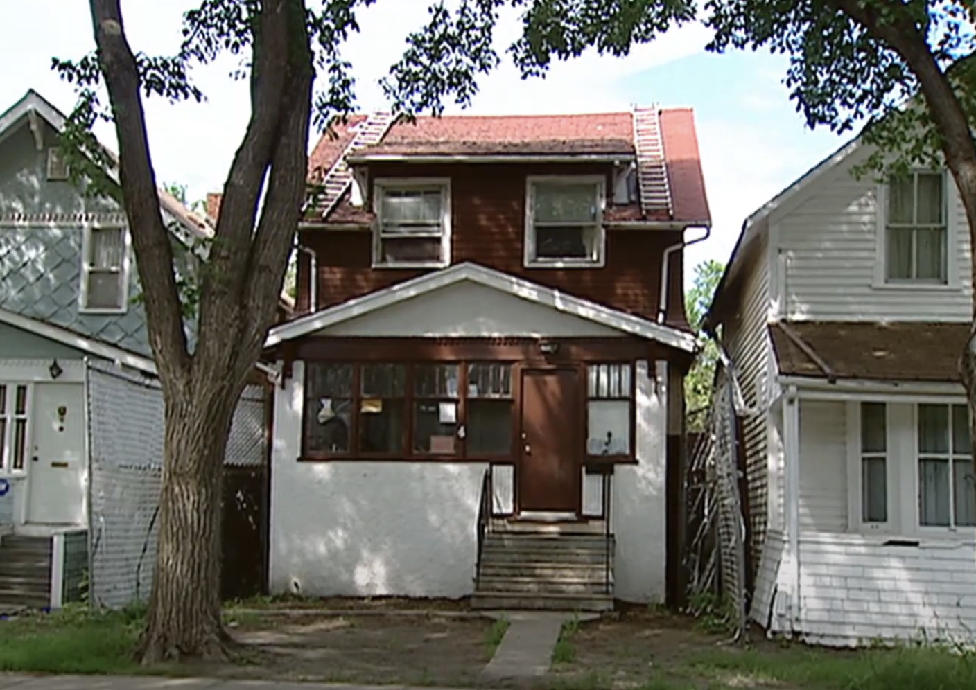 A two-story house with a brown upper level and a white lower level, flanked by two neighboring houses, with a tree in the front yard and a small walkway leading to the entrance