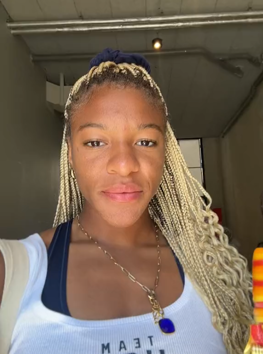 Ariana Ramsey posing for a selfie, wearing a tank top and a necklace; she has long braided hair tied back and is holding an orange item in her hand