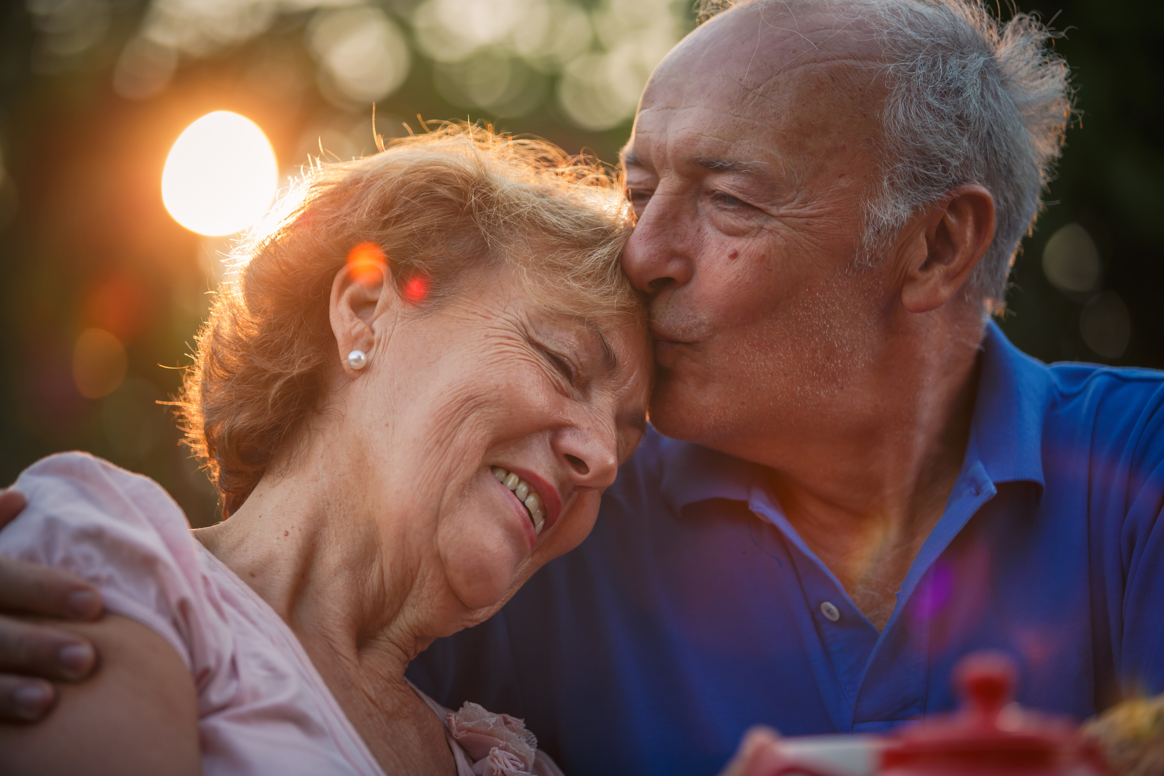 An elderly man kisses an elderly woman on the forehead as they enjoy a tender moment outdoors at sunset