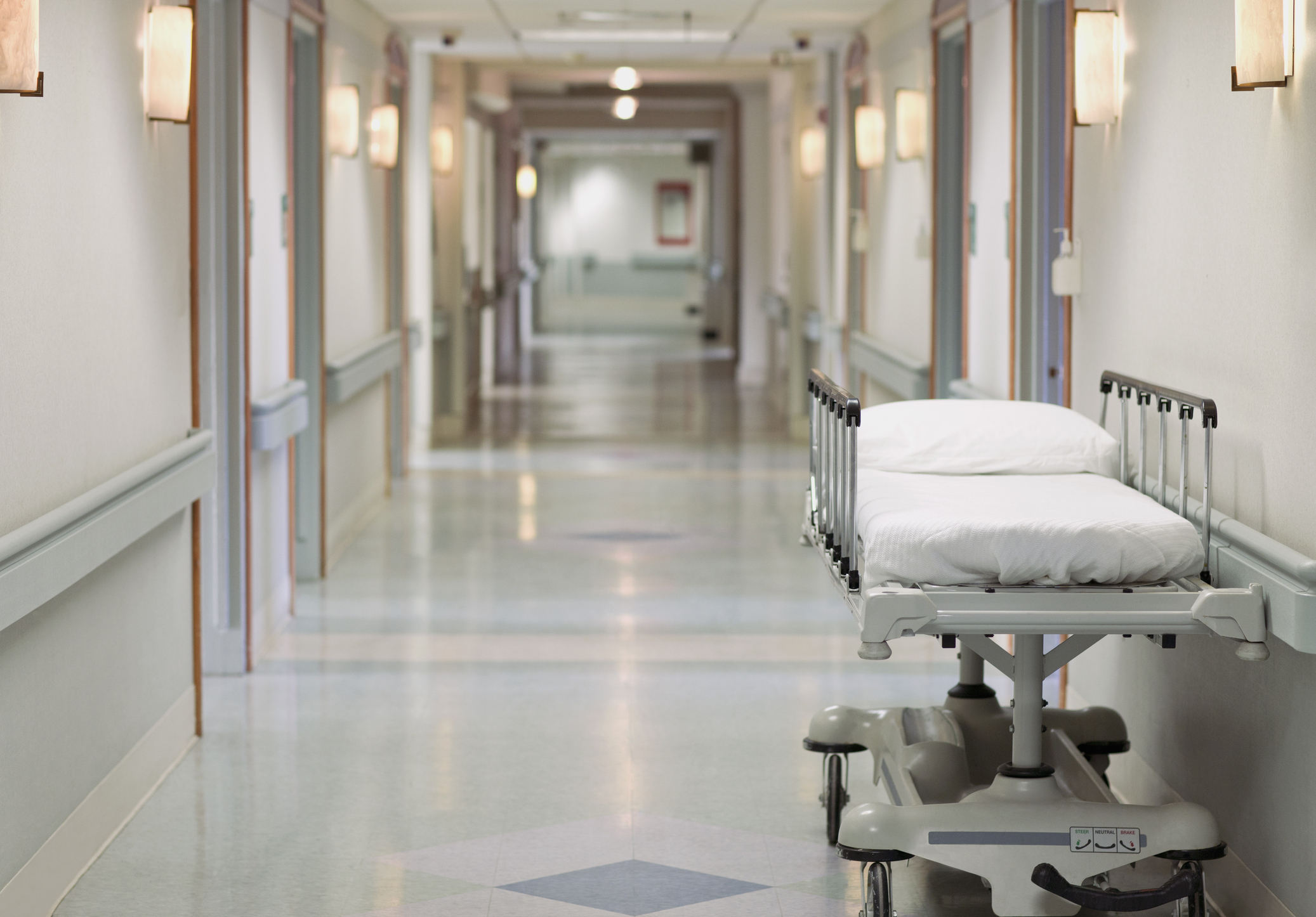 An empty hospital hallway with a white bed on wheels placed to the side. The hall is well-lit and appears clean and sterile