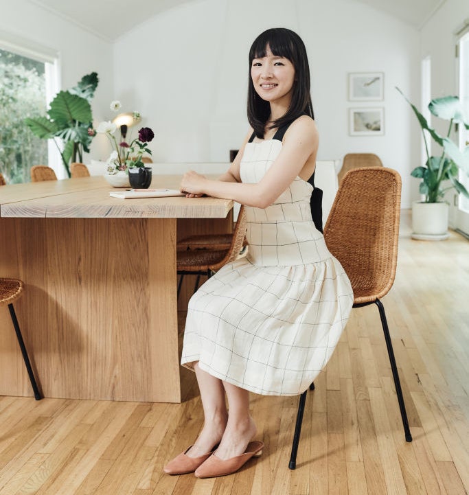 Marie Kondo sits at a wooden table in a minimalist room with large green plants and framed pictures on the walls