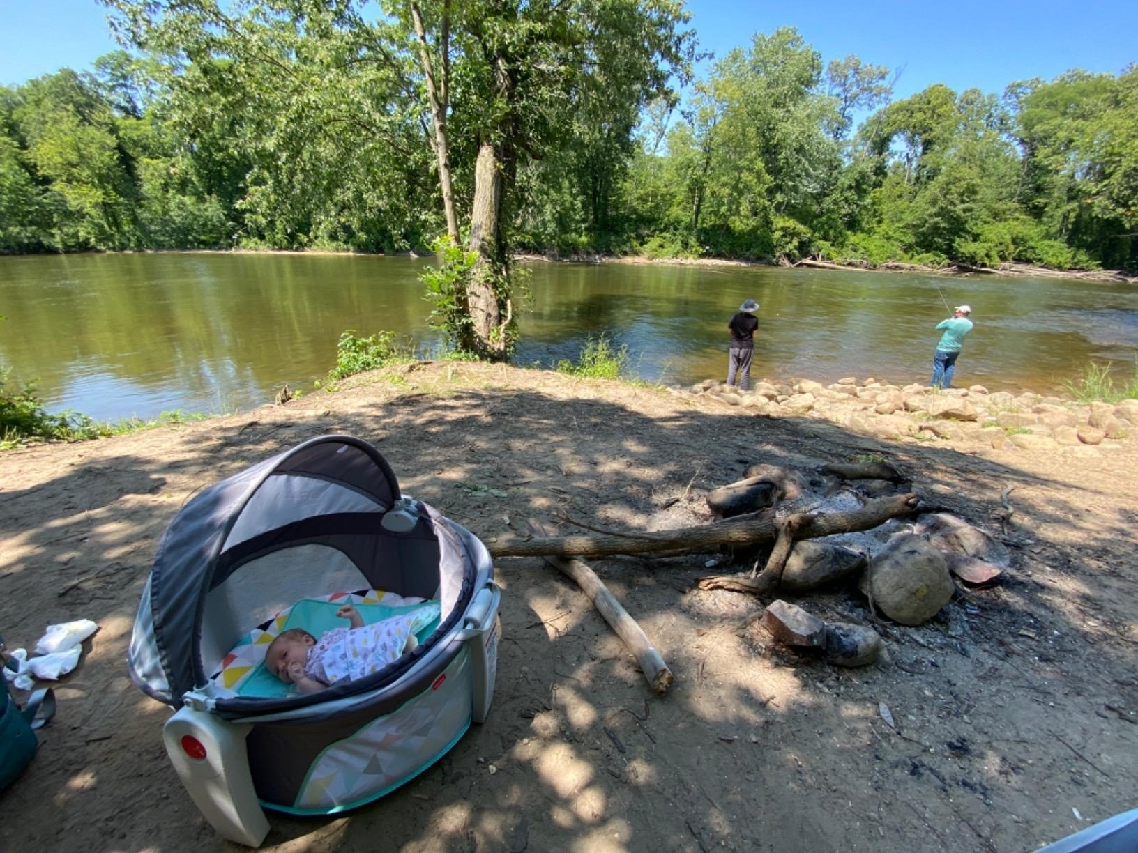 A baby sleeps in a portable crib by a riverside campsite with two people fishing in the background. The area is surrounded by trees