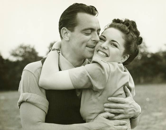 A man and woman in vintage-style attire share an affectionate embrace outdoors, both smiling happily