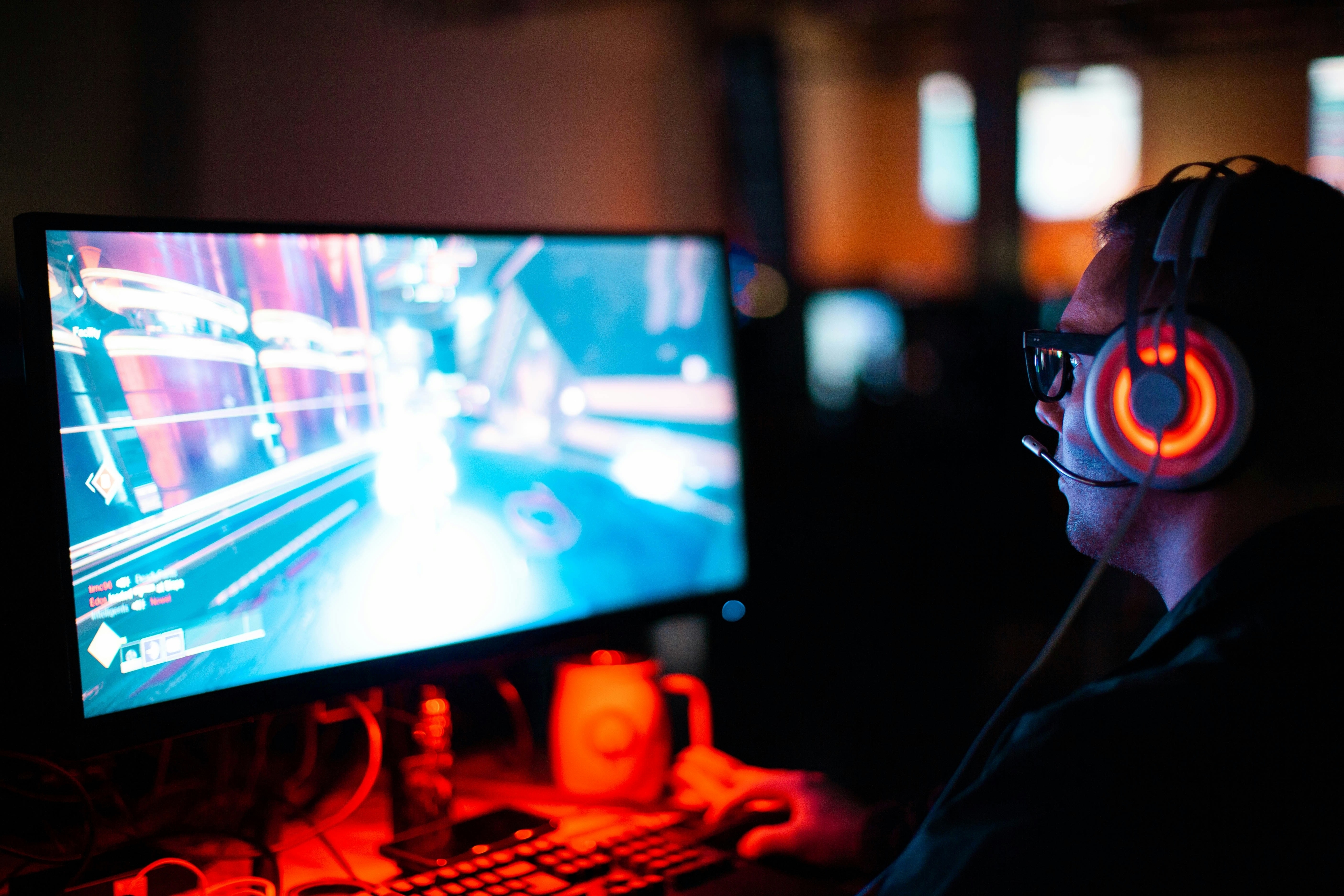 Person wearing headphones and glasses, gaming on a computer with a lit monitor screen showing a fast-paced game