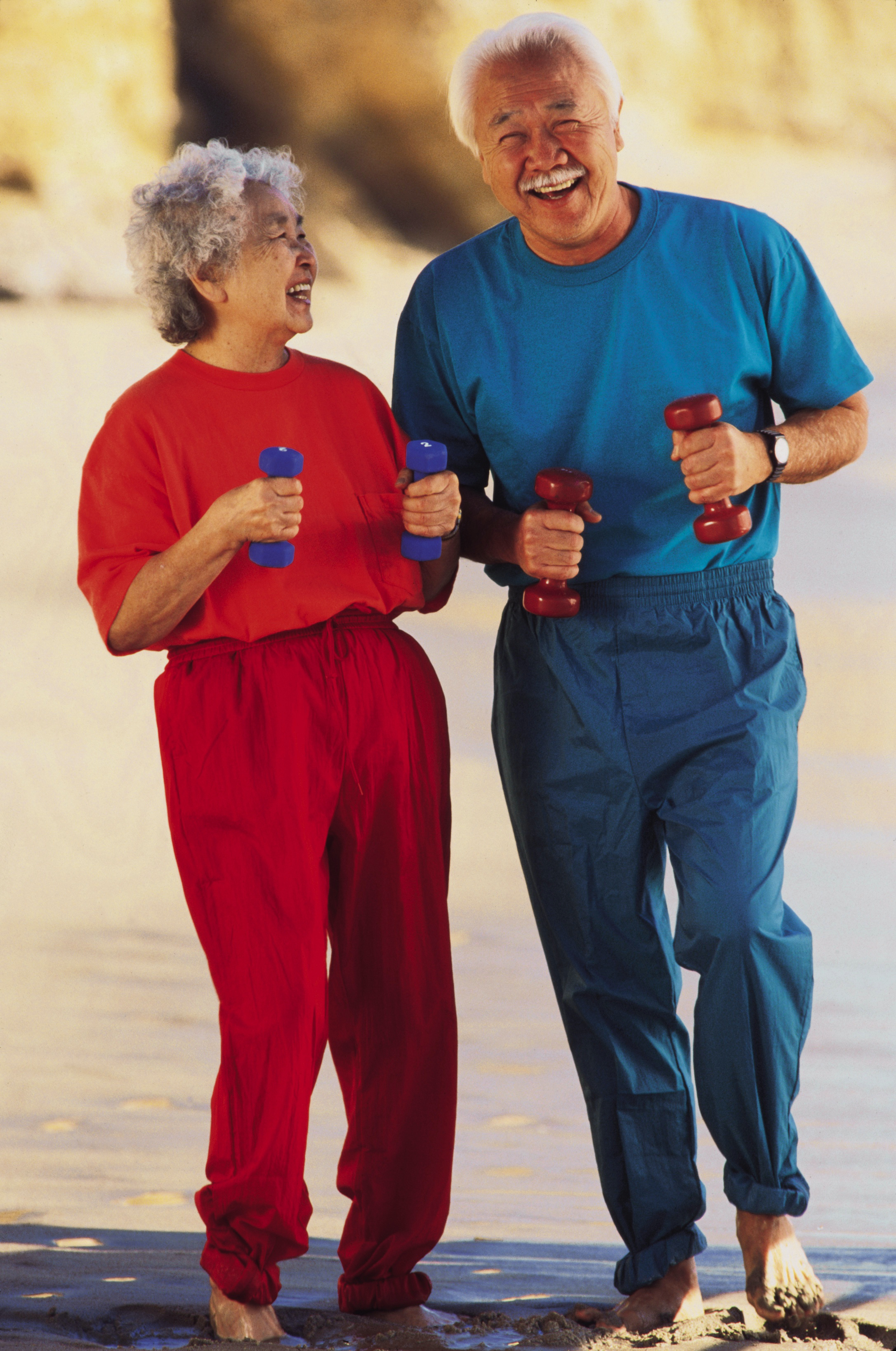 Elderly couple laughing and holding dumbbells on a beach, both wearing activewear