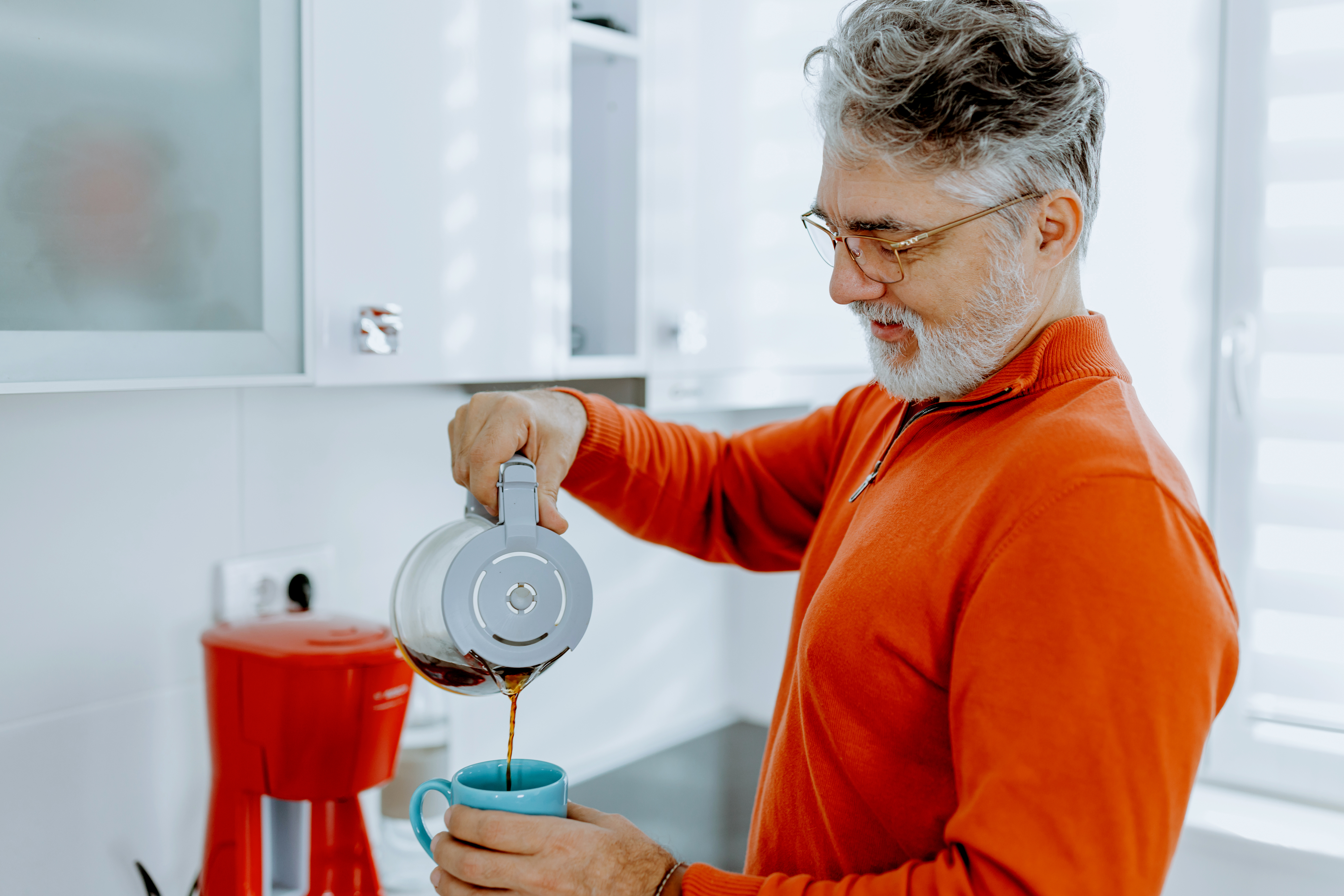 An older man pours coffee from a French press into a mug in a modern kitchen. He is smiling and wearing glasses and a long-sleeve shirt