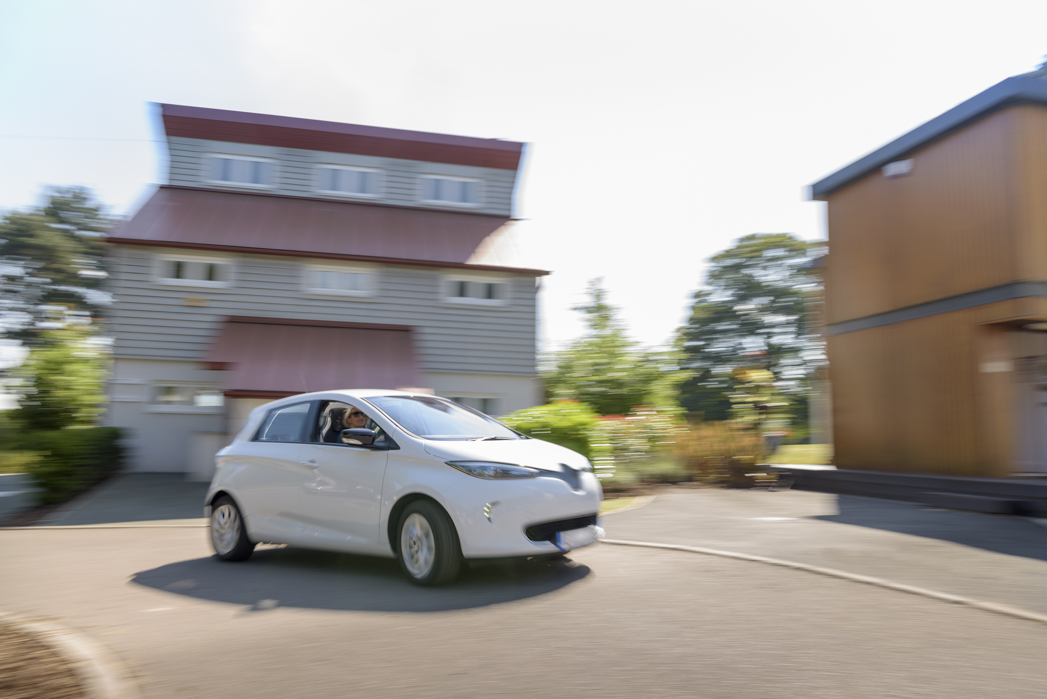 A person driving a small white electric car takes a turn in a residential area with a modern multi-story house and greenery in the background