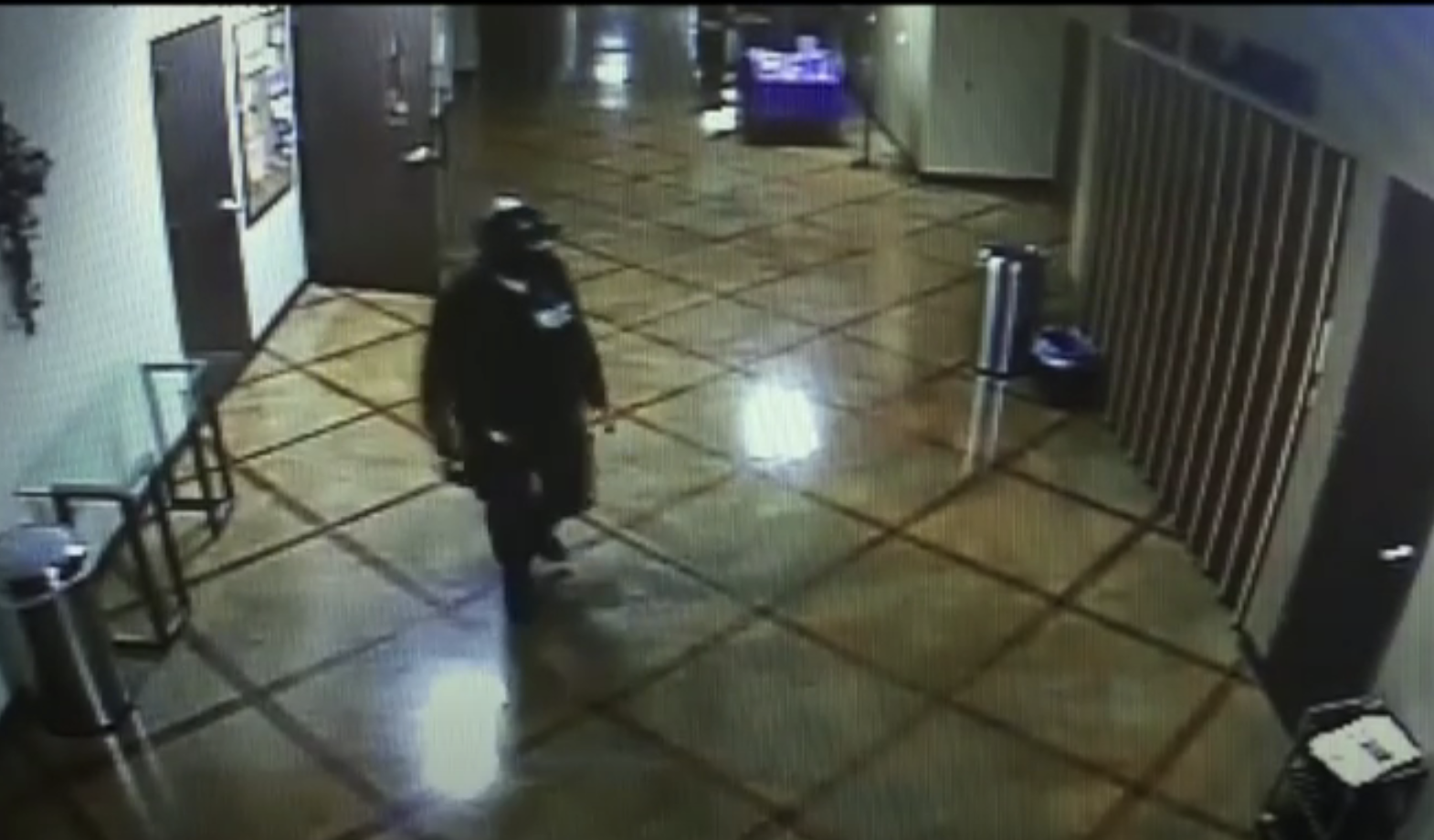 A surveillance camera captures an individual in dark clothing and helmet walking through a tiled hallway with trash cans and a bulletin board