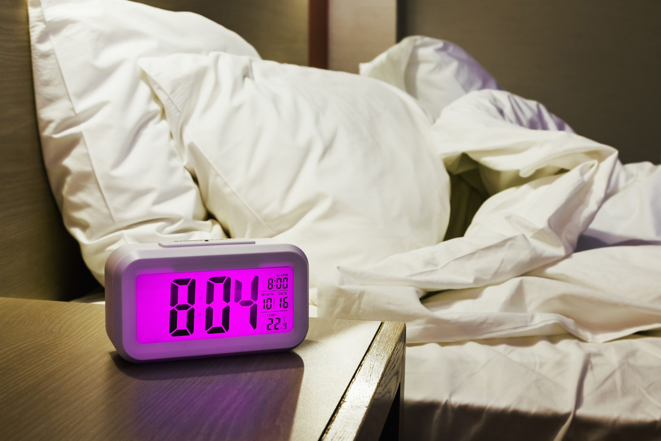 Digital alarm clock showing 8:04 on a nightstand next to an unmade bed with white pillows and blankets