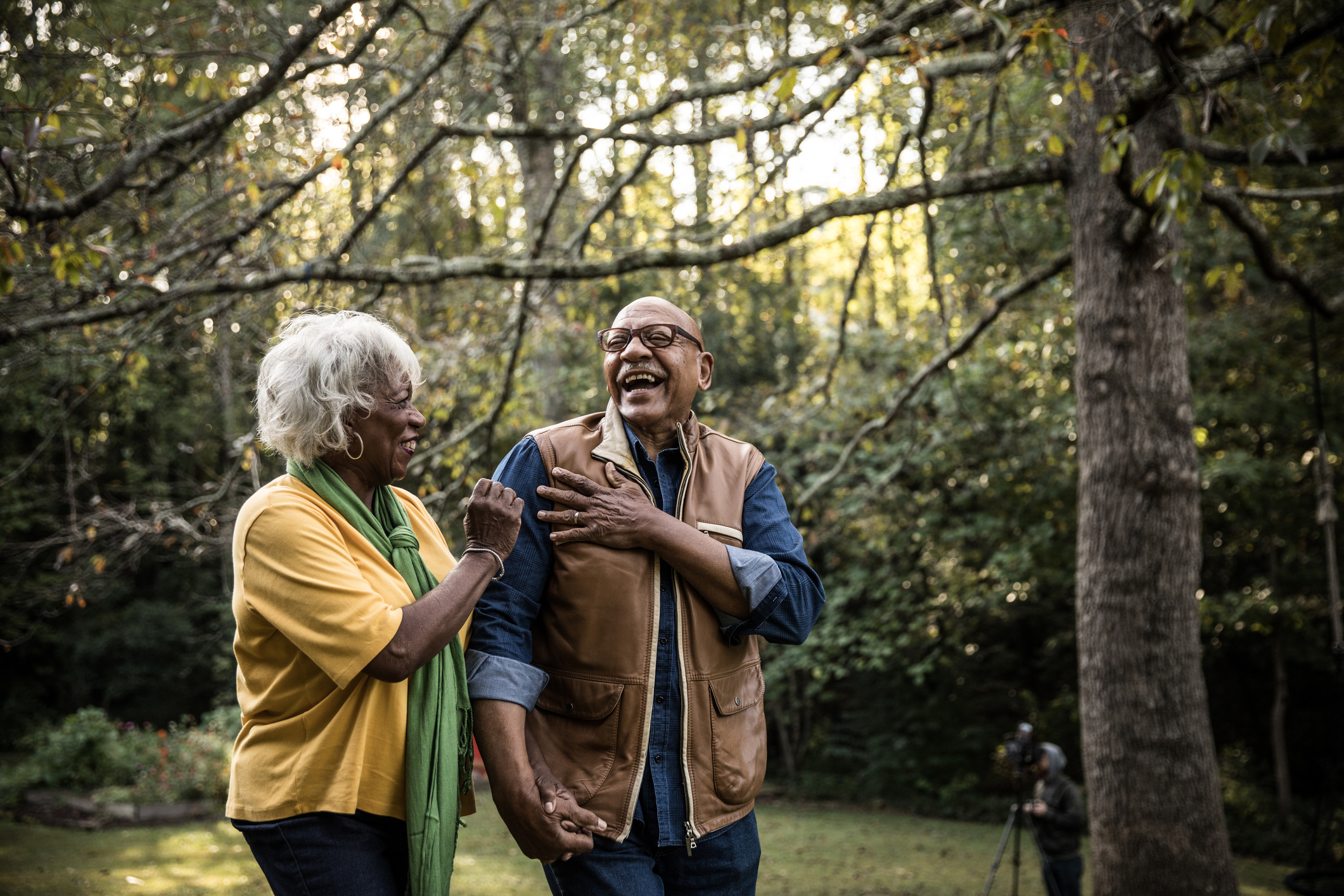 An elderly couple stands in a wooded area, laughing and holding hands. The woman wears a yellow top with a green scarf, and the man wears a tan vest over a blue shirt