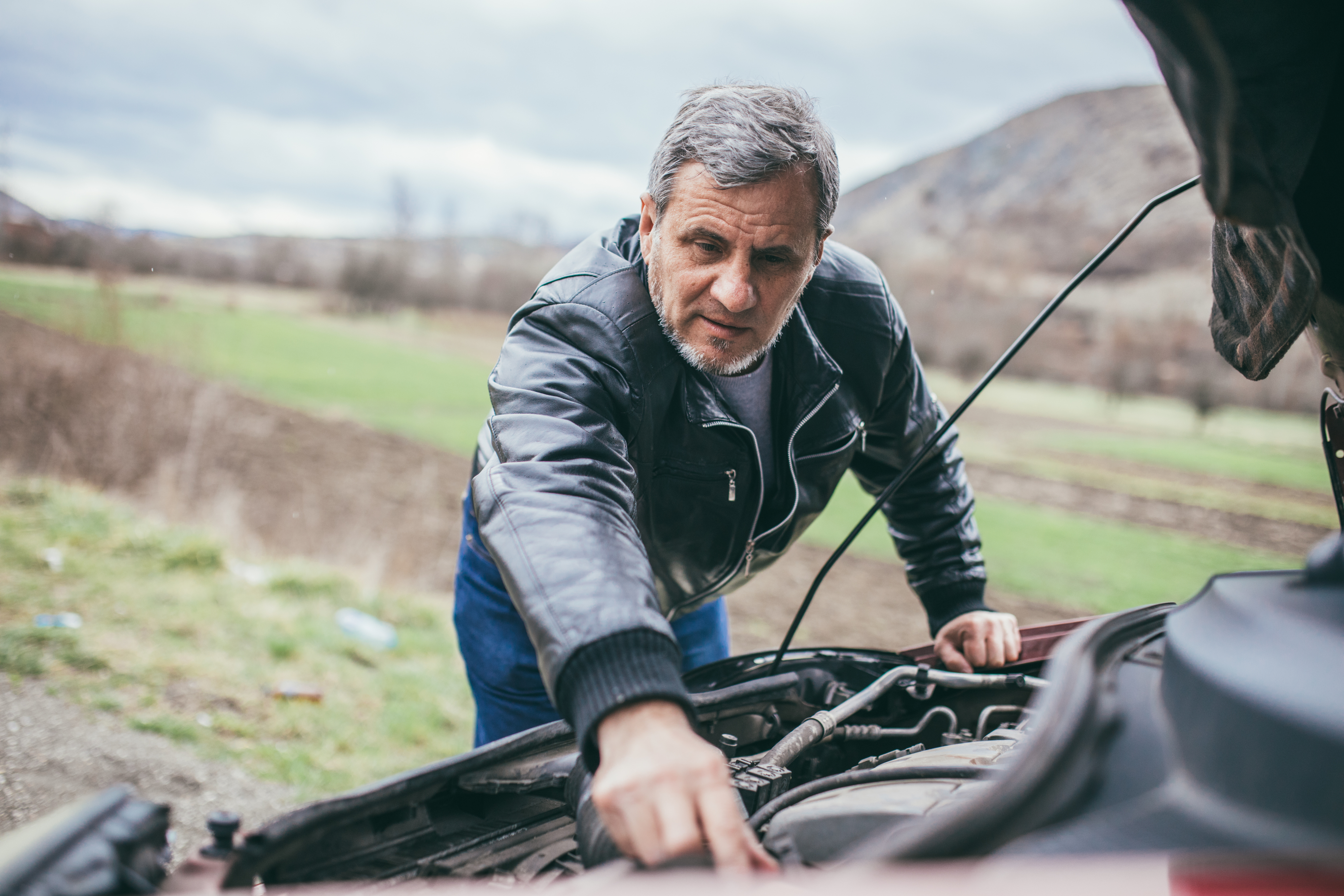 A man in a leather jacket looks under the hood of a car in a rural area. He is focused on inspecting something in the engine