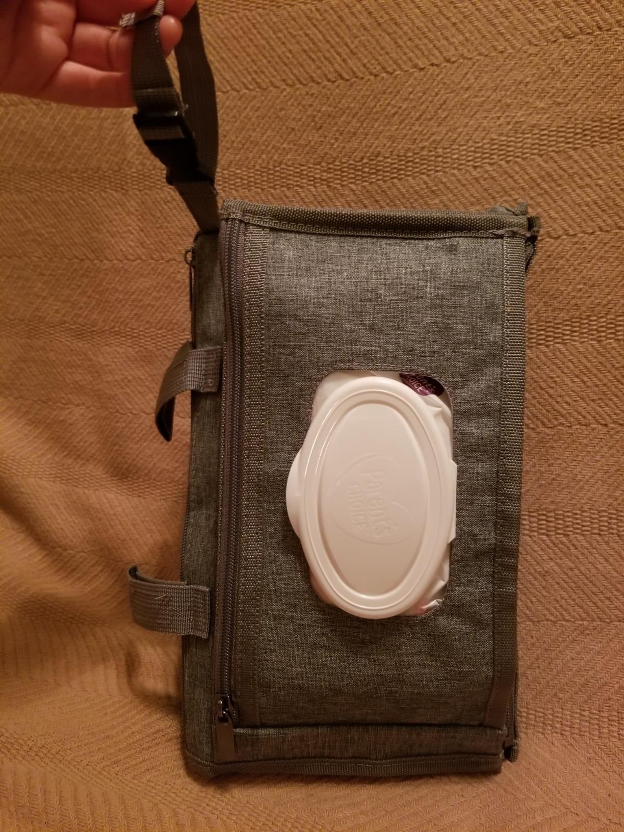 A manus  holds a rectangular cloth  container  with a zippered pouch  and a built-in, achromatic  integrative  hitch   dispenser