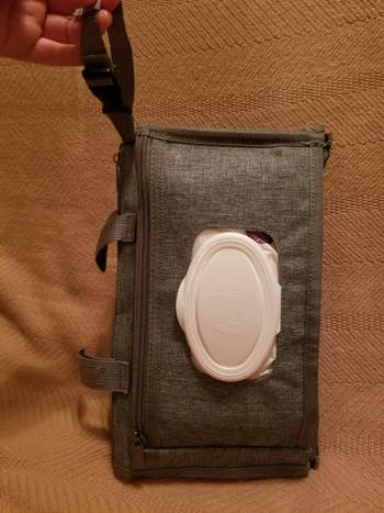 A manus holds a rectangular cloth container with a zippered pouch and a built-in, achromatic integrative hitch dispenser