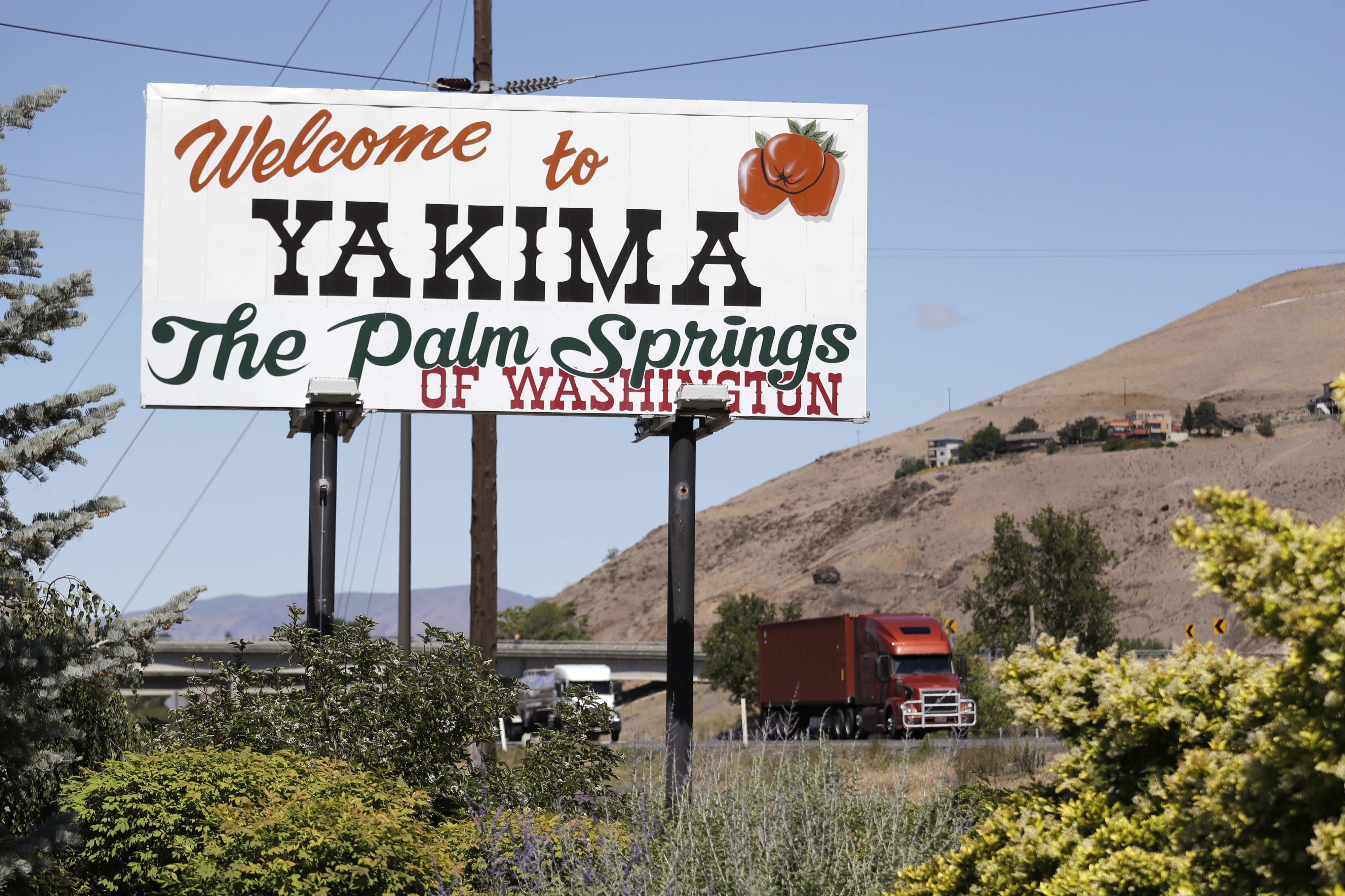 A roadside sign reads "Welcome to Yakima, The Palm Springs of Washington" with an image of a tomato. A red truck drives on a highway in the background